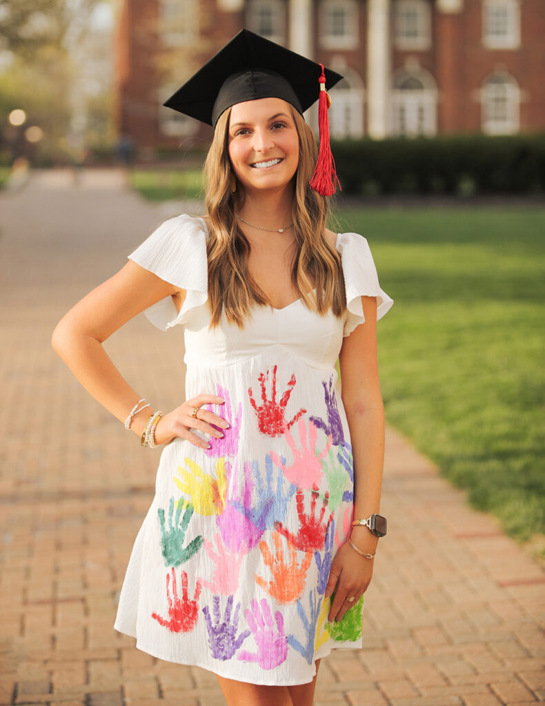An elementary education graduate smiling in a black cap with a red tassel, wearing a white dress decorated with colorful student handprints.