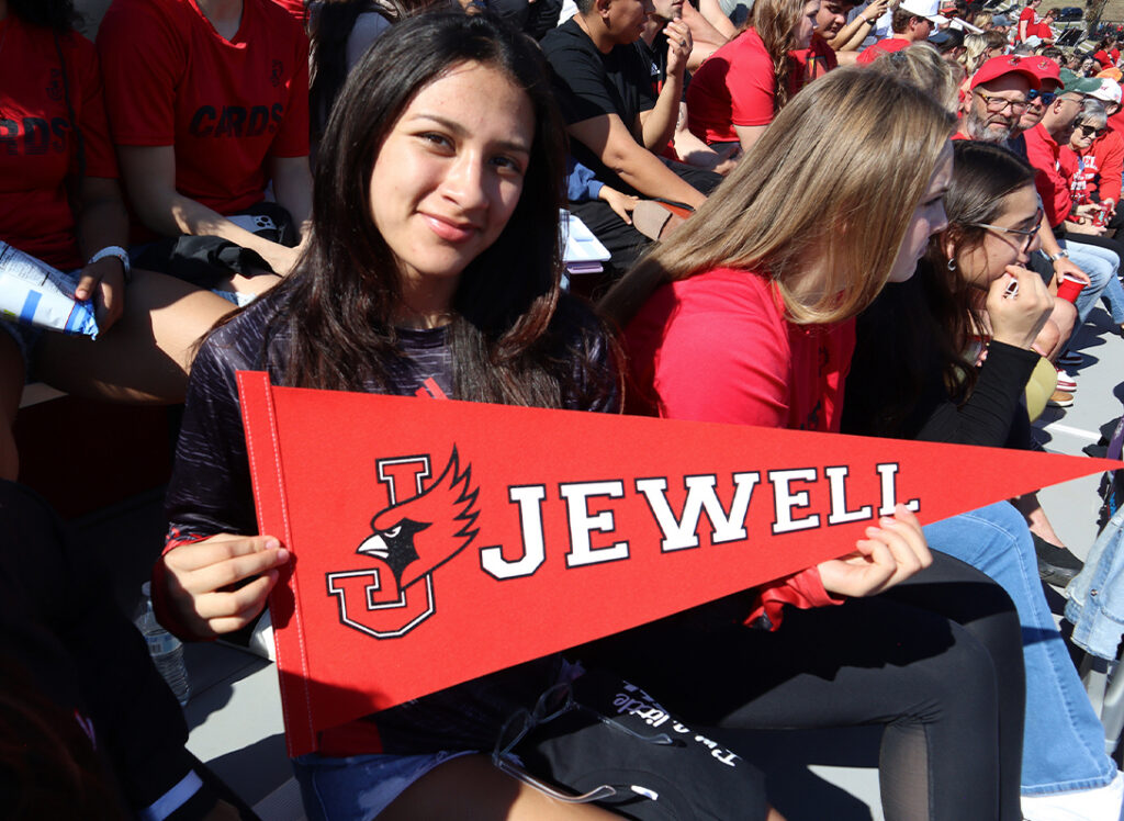A smiling female student sitting in stadium grandstands holding a red "Jewell" felt pennant.