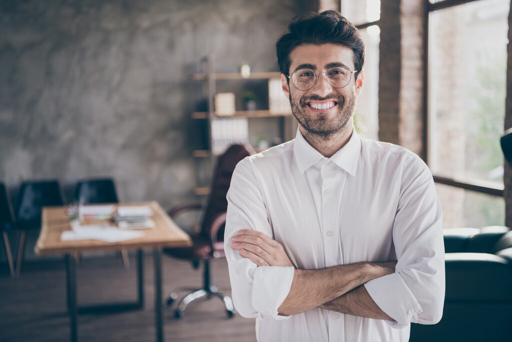A smiling male professional wearing round glasses and a white button-down shirt standing with arms crossed in an office environment.