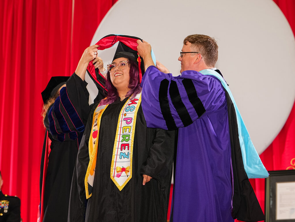 A female master's degree candidate receiving her academic hood during a graduation ceremony on stage.