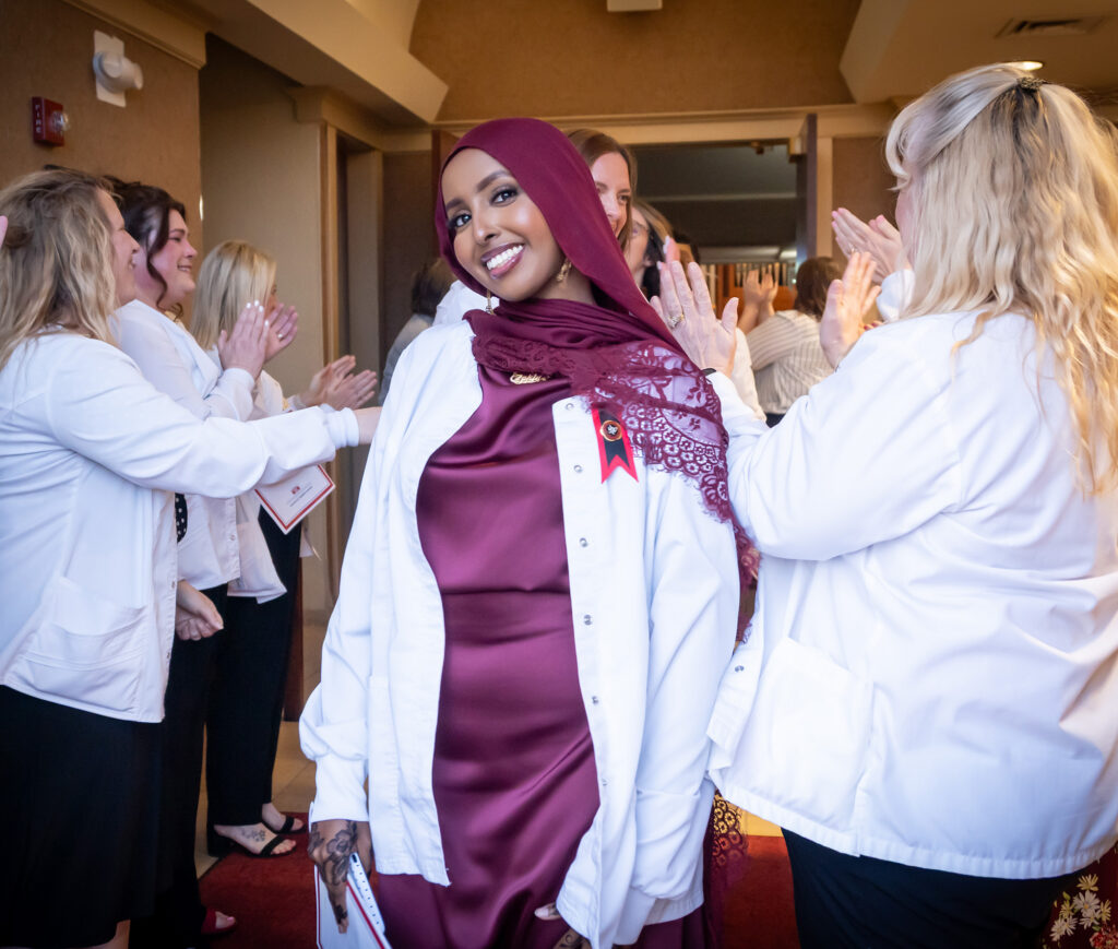 A nursing graduate wearing a maroon dress, matching hijab, and white lab coat smiling as peers applaud during a pinning ceremony.