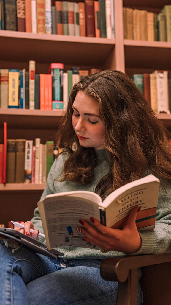 A student sitting in an armchair reading a book titled "Falling Into Theory" in front of library bookshelves.
