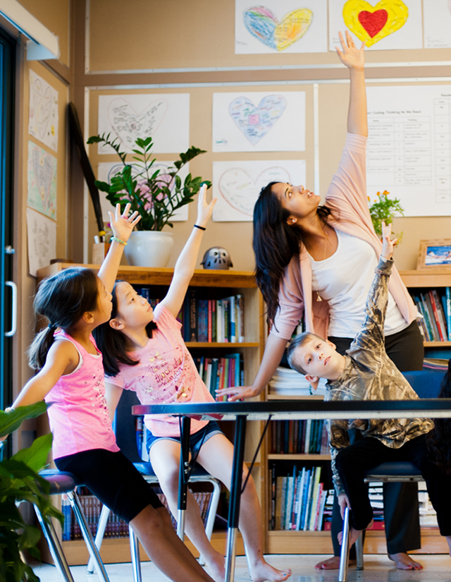 A teacher and young elementary students reaching upward together in a classroom stretch exercise.