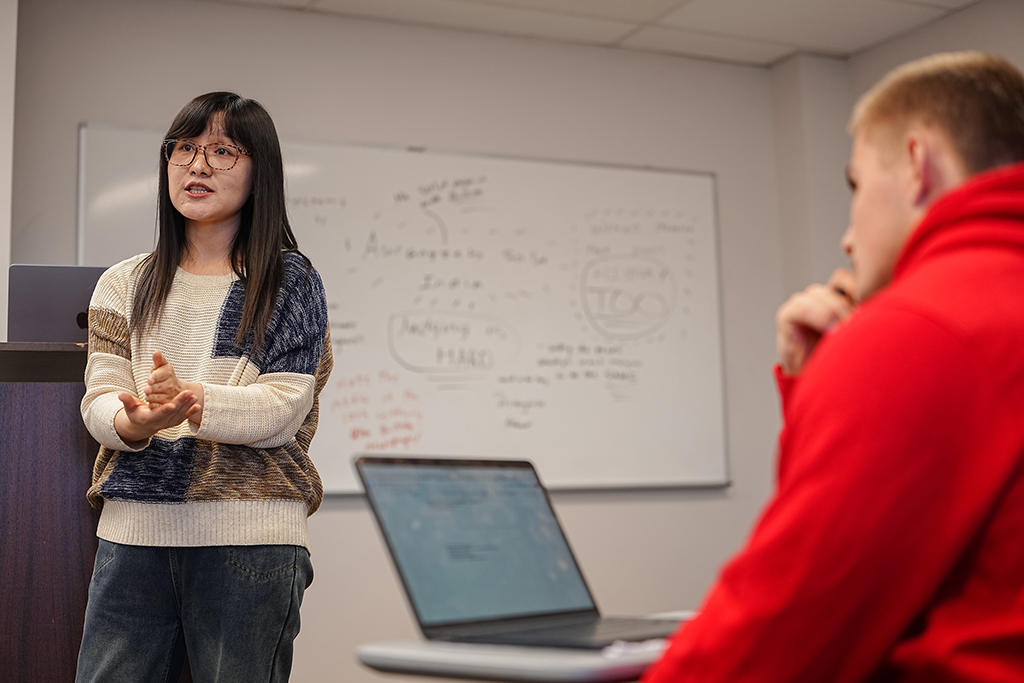Ge Zhu, Assistant Professor of Communication, speaking to a student wearing a red hoodie during a classroom discussion.