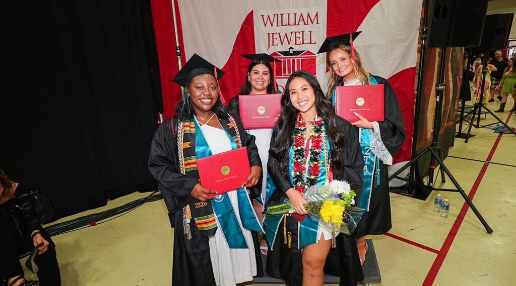 4 female graduates smiling and holding diploma covers while posing in caps and gowns in front of a William Jewell backdrop.