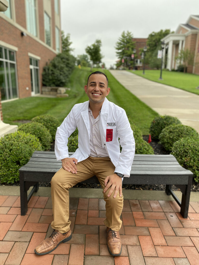 Manny Macias, a nursing student at William Jewell College wearing a white lab coat with a red nursing ribbon, smiling while seated on an outdoor bench.