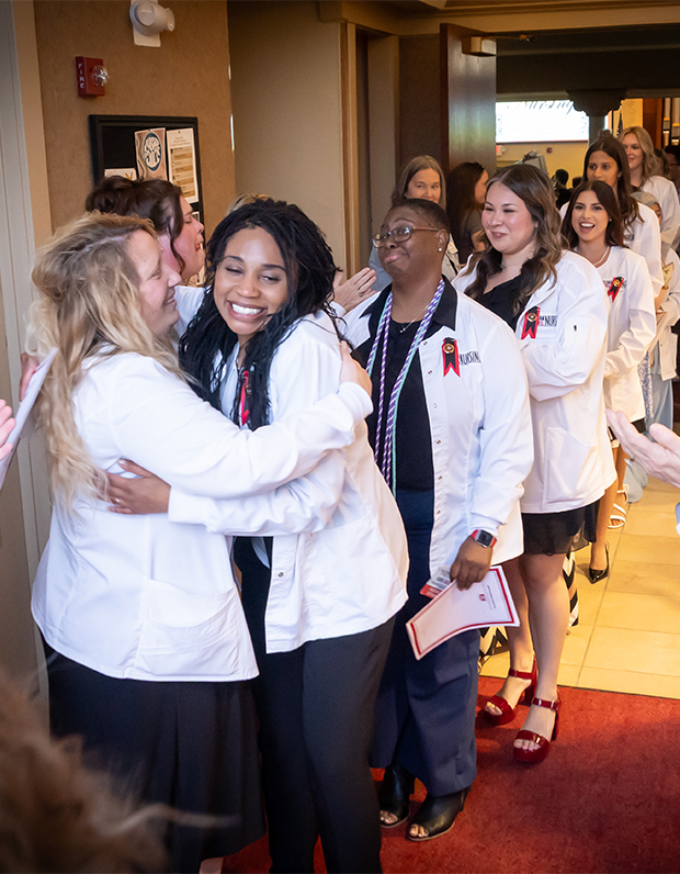 Nursing graduates in white lab coats hugging and celebrating as supporters cheer during a pinning ceremony.
