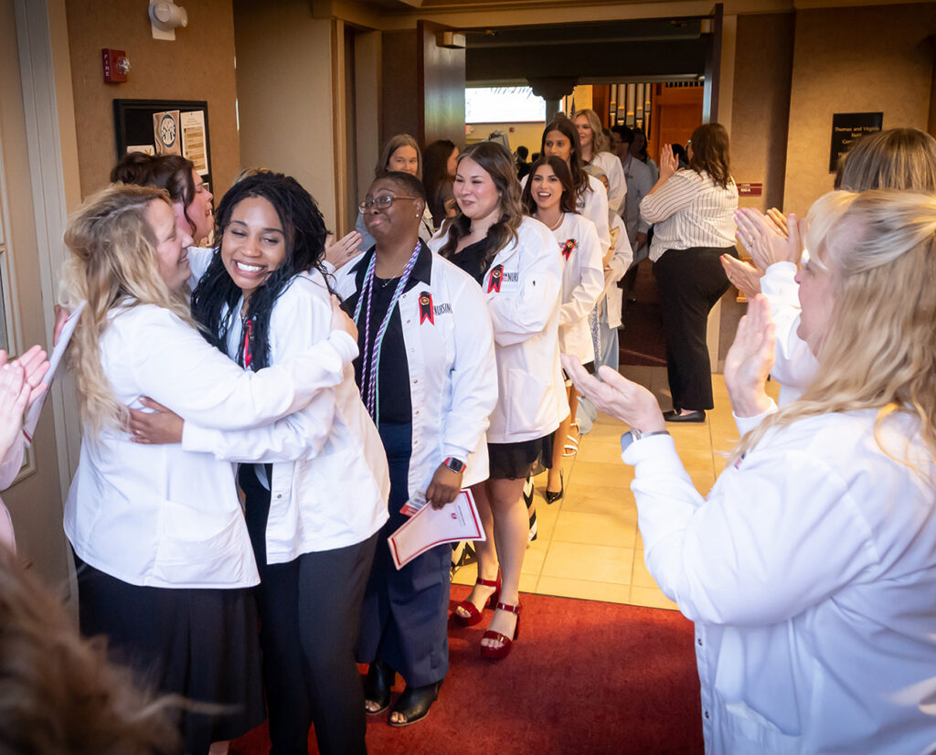 Nursing graduates in white lab coats hugging and celebrating as supporters cheer during a pinning ceremony.