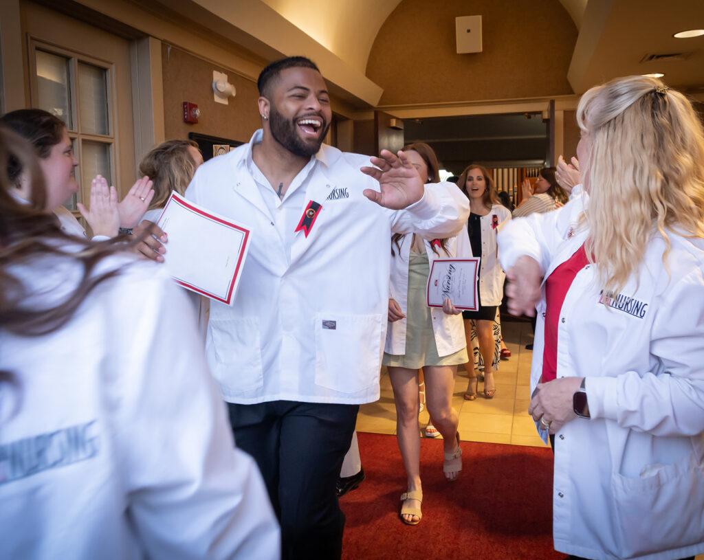 A smiling male nursing graduate high-fiving supporters as he walks through a line of peers during a pinning ceremony.