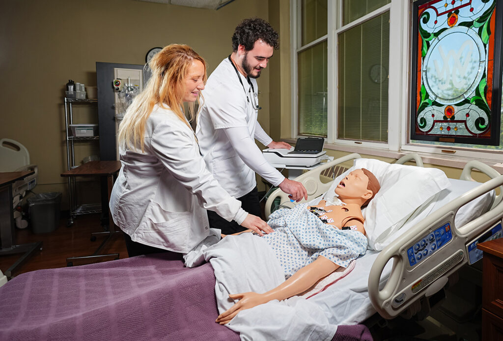 A nursing instructor and student practicing medical procedures on a patient simulator mannequin in a training lab.
