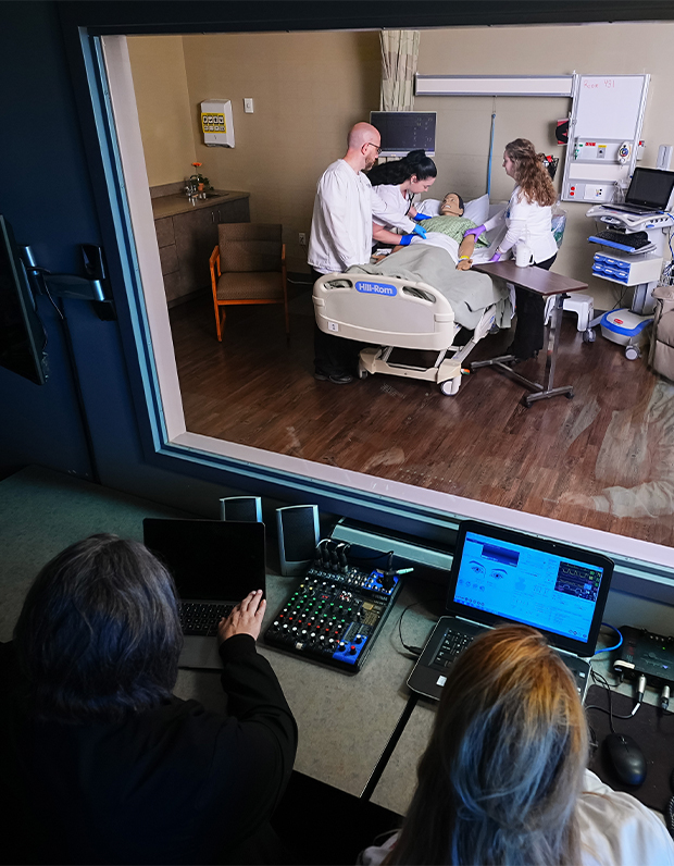 View through an observation window of nursing students practicing clinical skills on a training mannequin while instructors monitor controls.