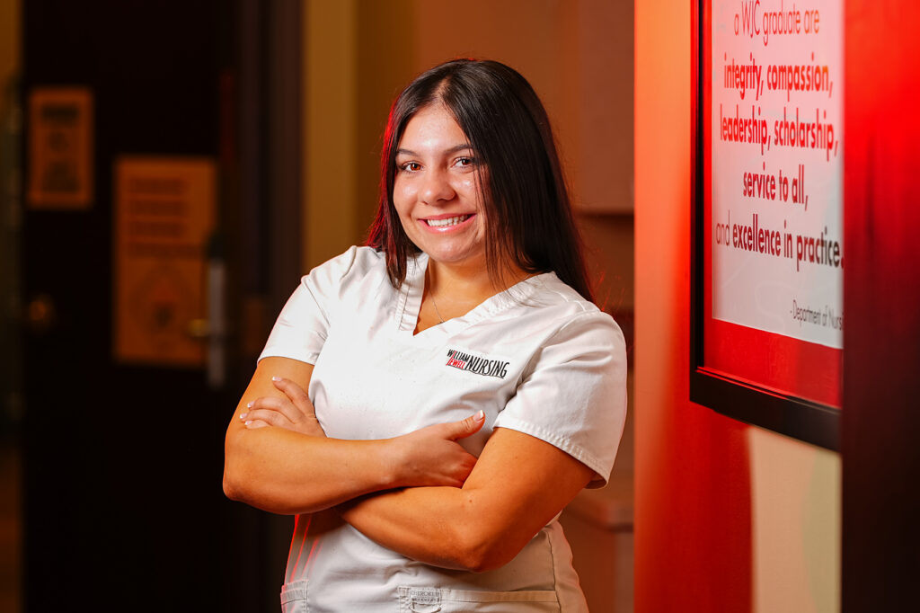 A female nursing student smiling with arms crossed in a white uniform scrub top.