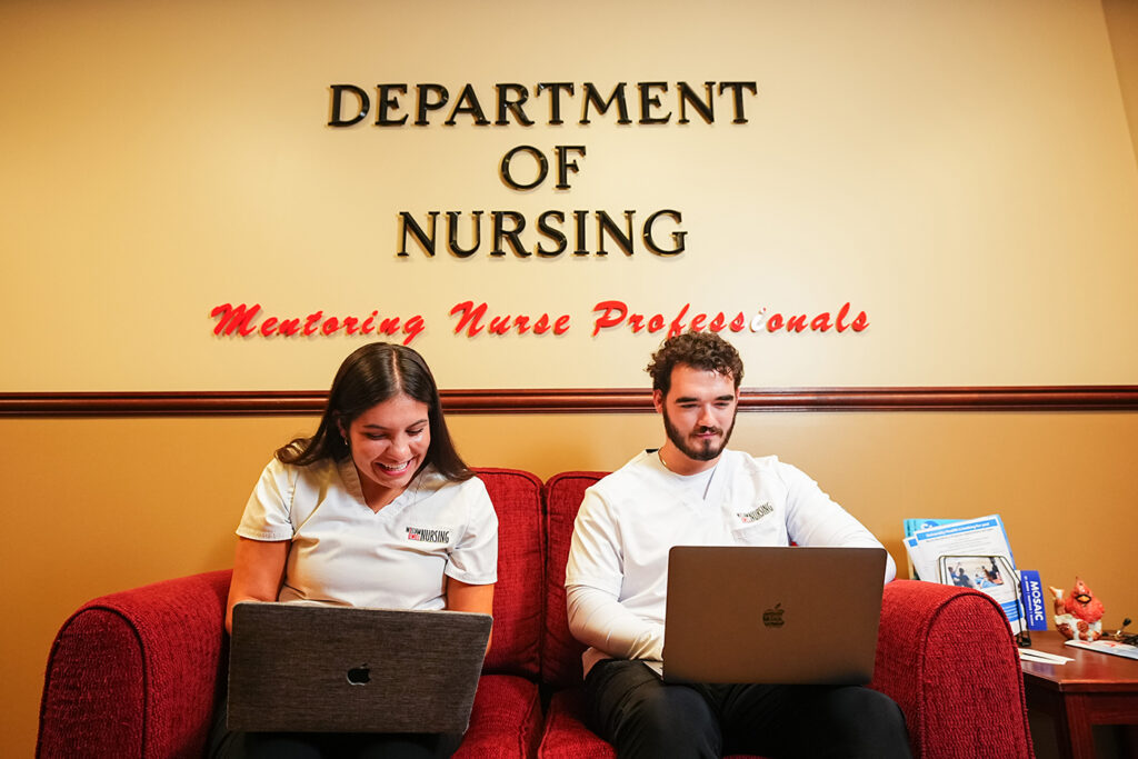 Two nursing students sitting on a red couch working on laptops under a "Department of Nursing" wall sign.