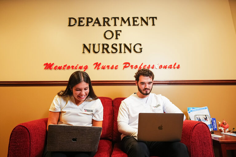 Two nursing students sitting on a red couch working on laptops under a "Department of Nursing" wall sign.