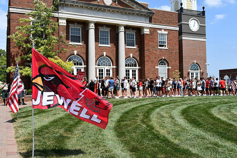 A line of incoming students walking across the lawn in front of John Gano Memorial Chapel during new student orientation, with a red Jewell flag in the foreground.