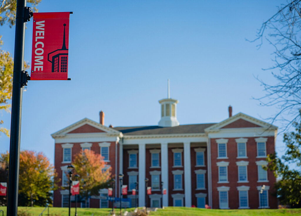 A red "Welcome" banner hanging on a lamp post with the historic brick Jewell Hall building blurred in the background.