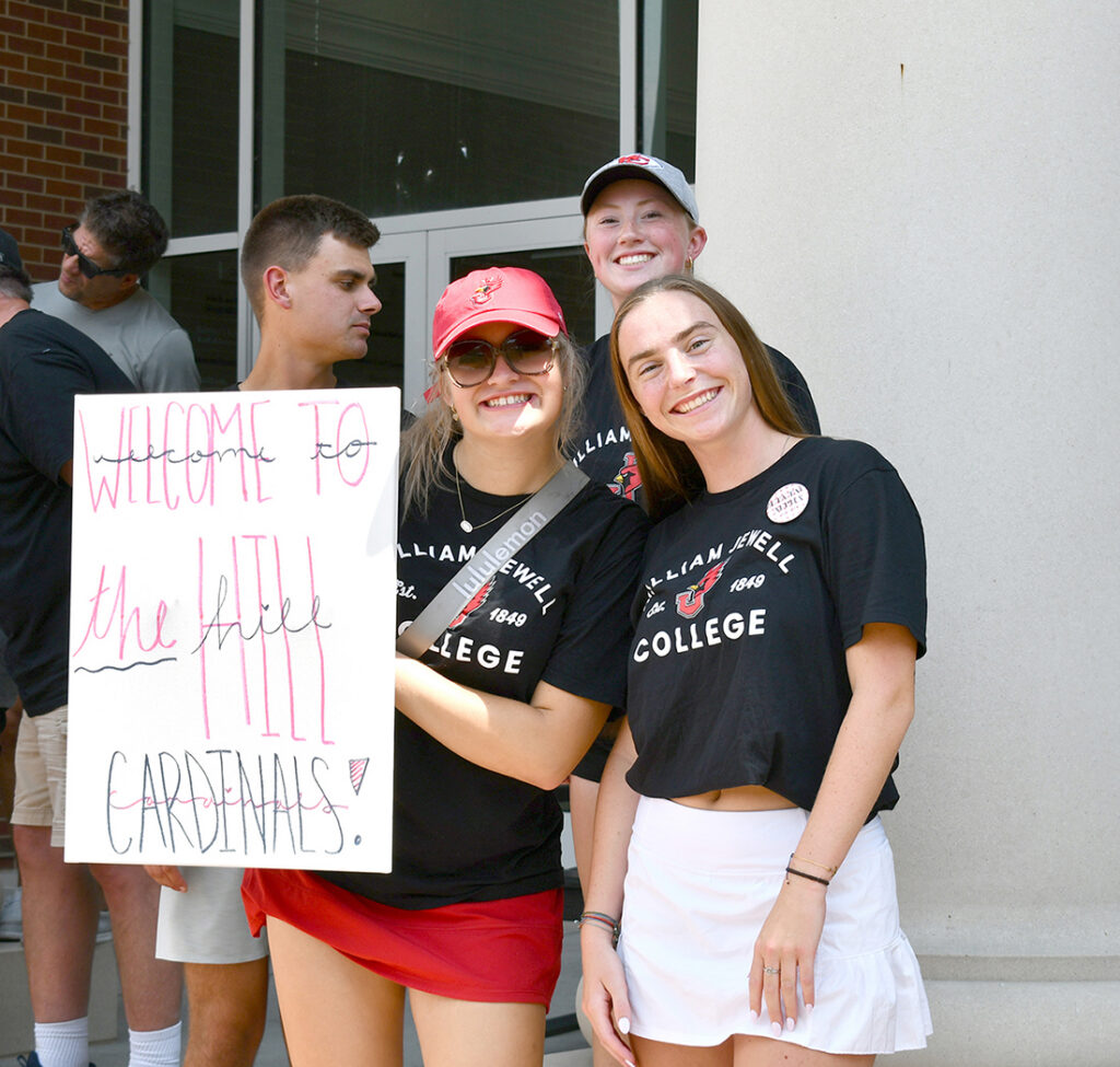 Students smiling and holding a handmade poster that reads "Welcome to the Hill Cardinals!" during a campus welcome event.