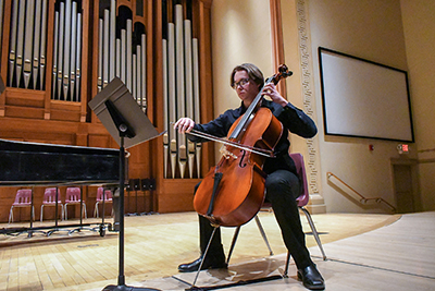 A student wearing glasses sitting on stage and playing a cello during a music recital in Gano Chapel.