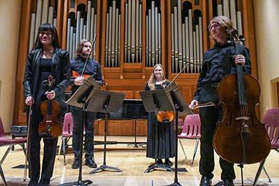 A student string quartet holding their instruments and smiling on stage in front of pipe organ pipes.