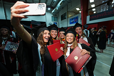 A group of graduates smiling and taking a selfie while holding their red diploma covers inside the fieldhouse.