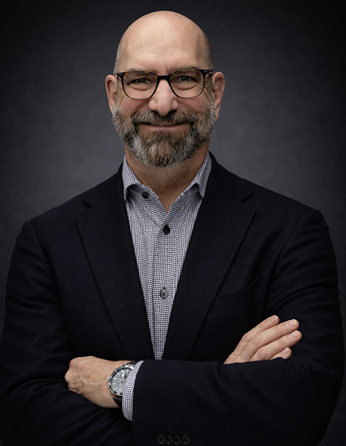 Headshot of man with crossed arms wearing glasses and a blazer.
