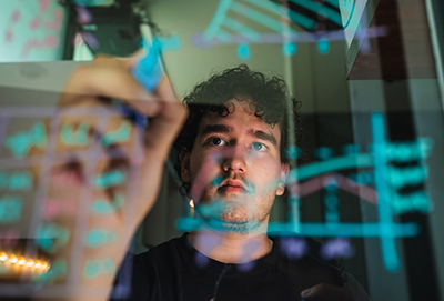 A student seen through a clear glass board drawing diagrams and writing equations with colorful dry-erase markers.