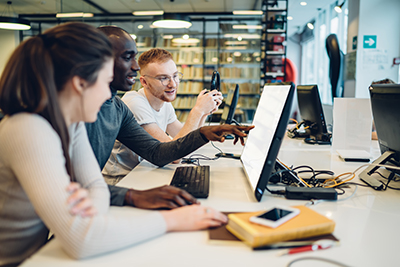 A group of students sitting at a computer workstation collaborating and pointing at a screen in a library.