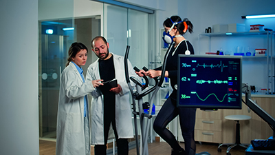 2 researchers in lab coats reviewing data on a tablet while a woman exercises on an elliptical trainer with monitoring equipment.