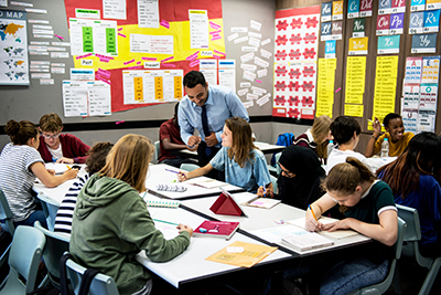 A teacher leaning down to assist students working at a shared table in a colorful classroom.