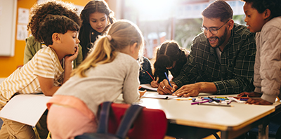 A male teacher assisting young elementary students seated around a table during an art or writing activity.