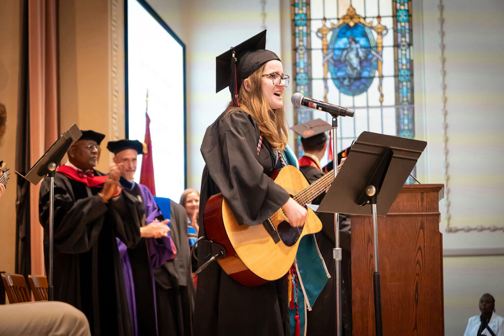 A graduating student playing acoustic guitar and singing at a microphone on stage during a Baccalaureate service in John Gano Memorial Chapel.