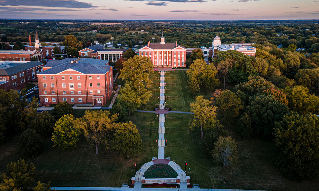 An aerial sunset view of the William Jewell College campus, featuring Jewell Hall, Quad walkways, surrounding academic buildings, and tree lines.