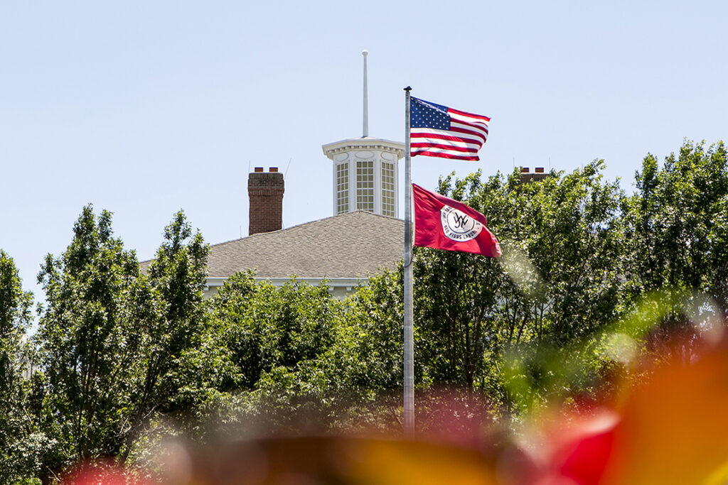 The American flag and William Jewell College flag flying on a flagpole with the cupola of Jewell Hall visible behind green trees.