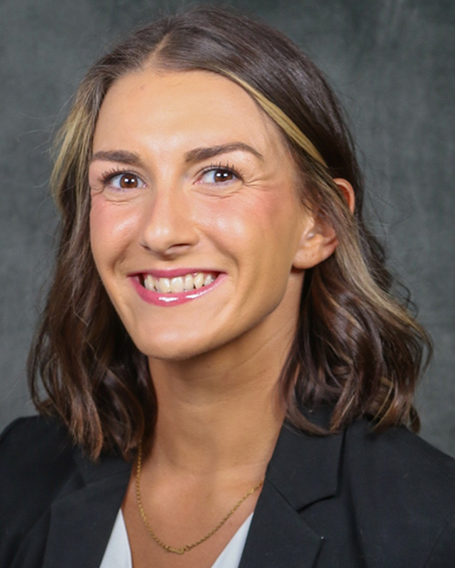 Jill Brown, Director of Athletic Performance, smiling in a dark suit, light blue shirt, and red-and-blue striped tie.