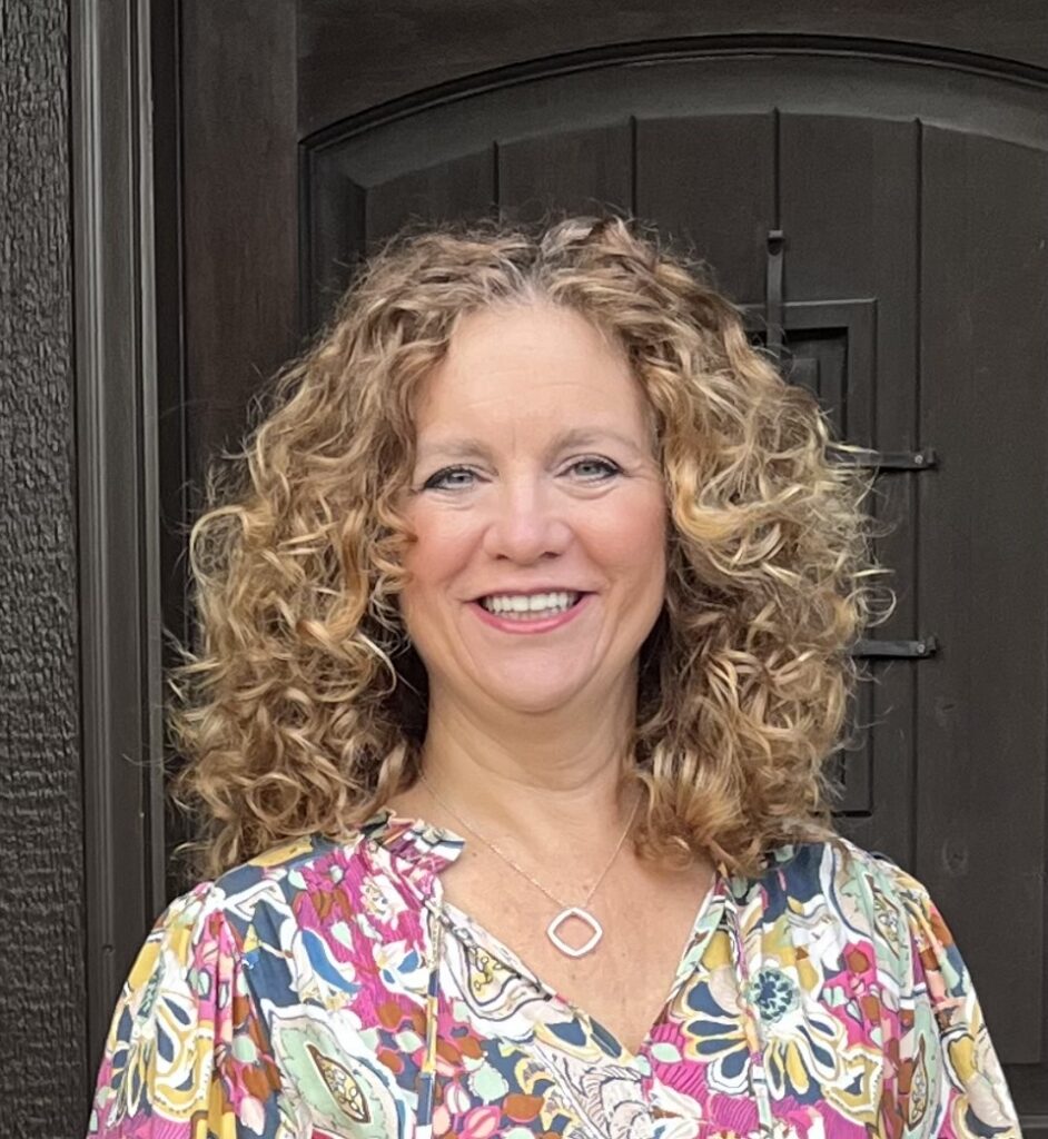 Dr. Jeanine Hastings, smiling with curly blonde hair and wearing a colorful floral-pattern top in front of a wooden door.