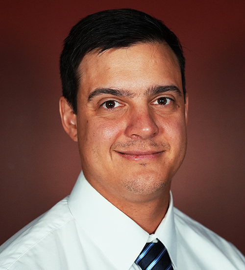 Dr. Alex Von Schulze, Assistant Professor at William Jewell College, smiling in a white dress shirt and striped tie against a dark red backdrop.