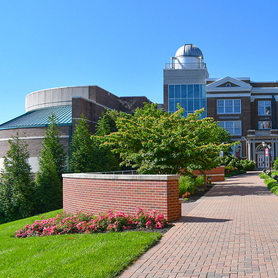 A brick walkway leading past landscaping toward the White Science Center observatory dome on a sunny day.
