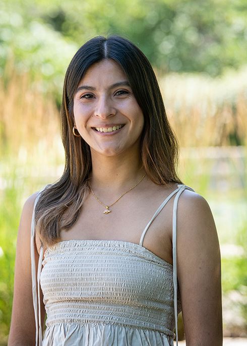 Erika Pinto, smiling outdoors with long dark hair, wearing a white smocked top and gold necklace.