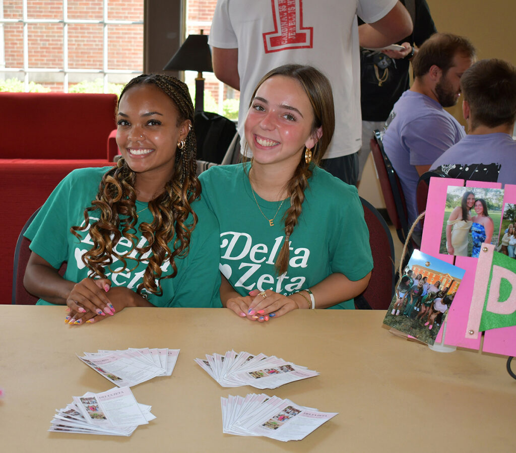 two women wearing green T-shirts seated at a table with information about their sorority