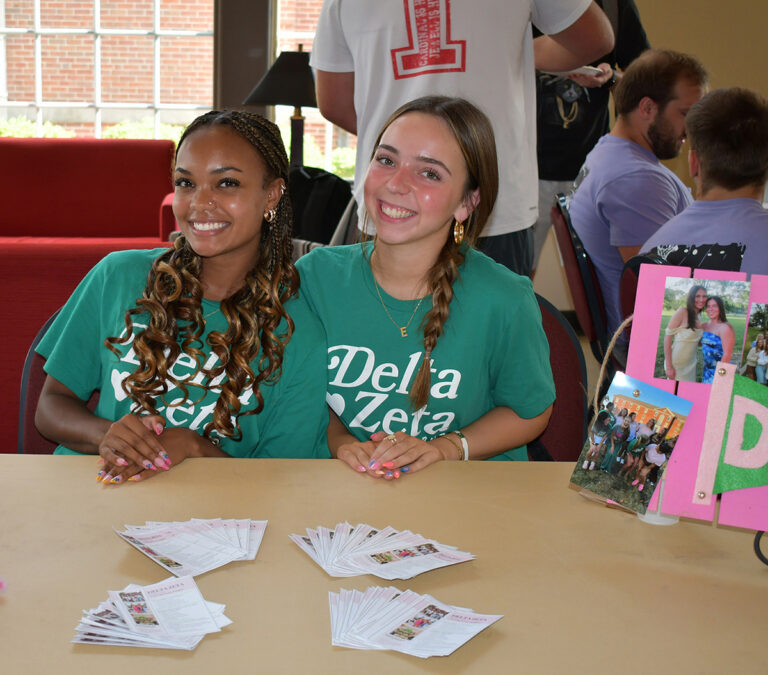 two women wearing green T-shirts seated at a table with information about their sorority