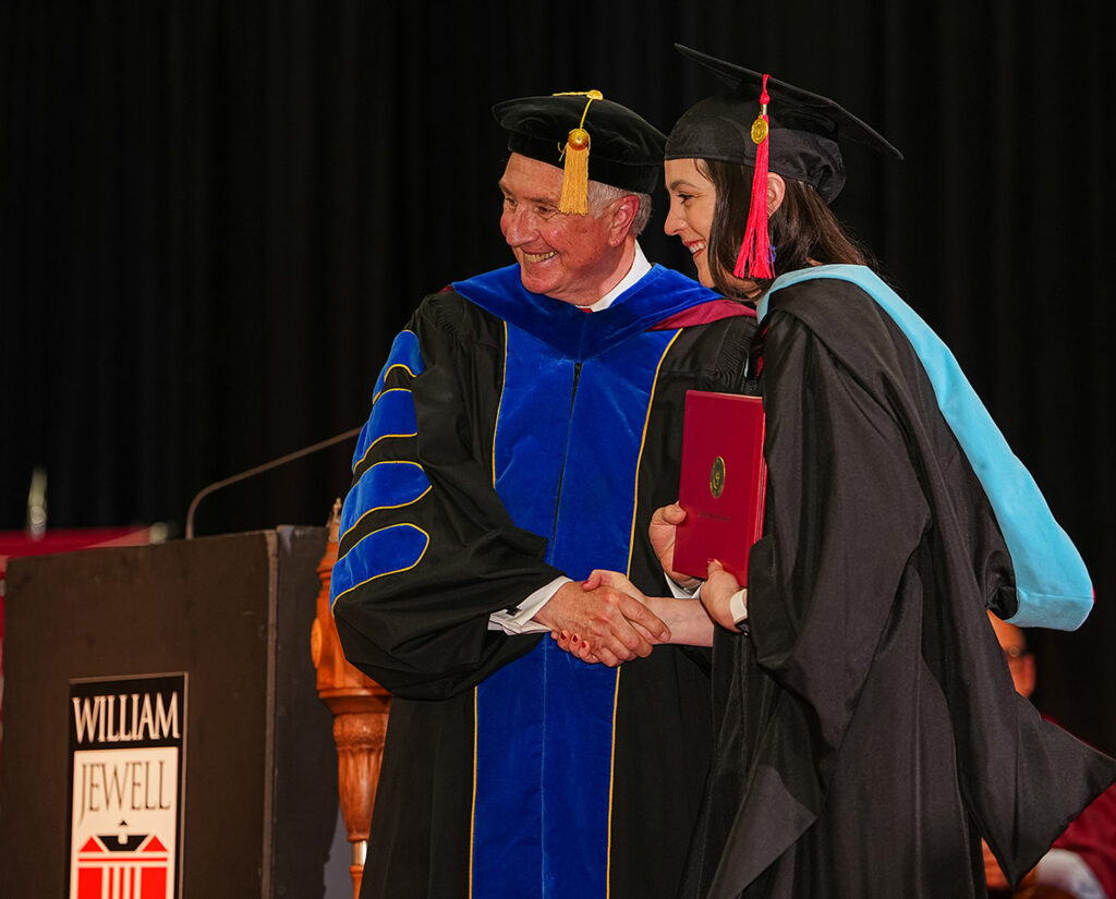woman wearing cap, gown, master's hood receiving diploma from president at commencement