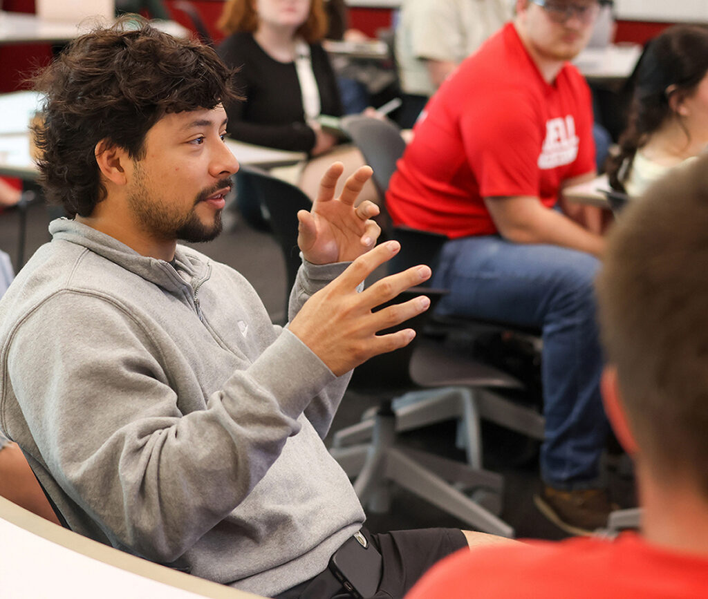 student seated in classroom, presenting a point to classmates