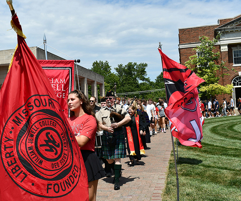 line of students walking around campus quad behind flag bearers and bagpipers