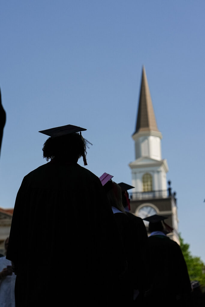 grads in caps and gowns walking single file past campus chapel
