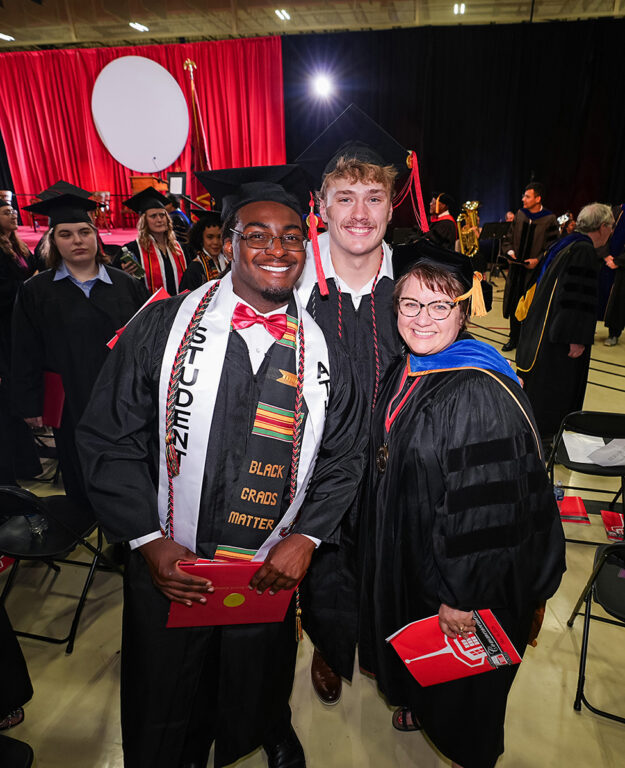 2 male grads posing with professor Tara Allen at commencement