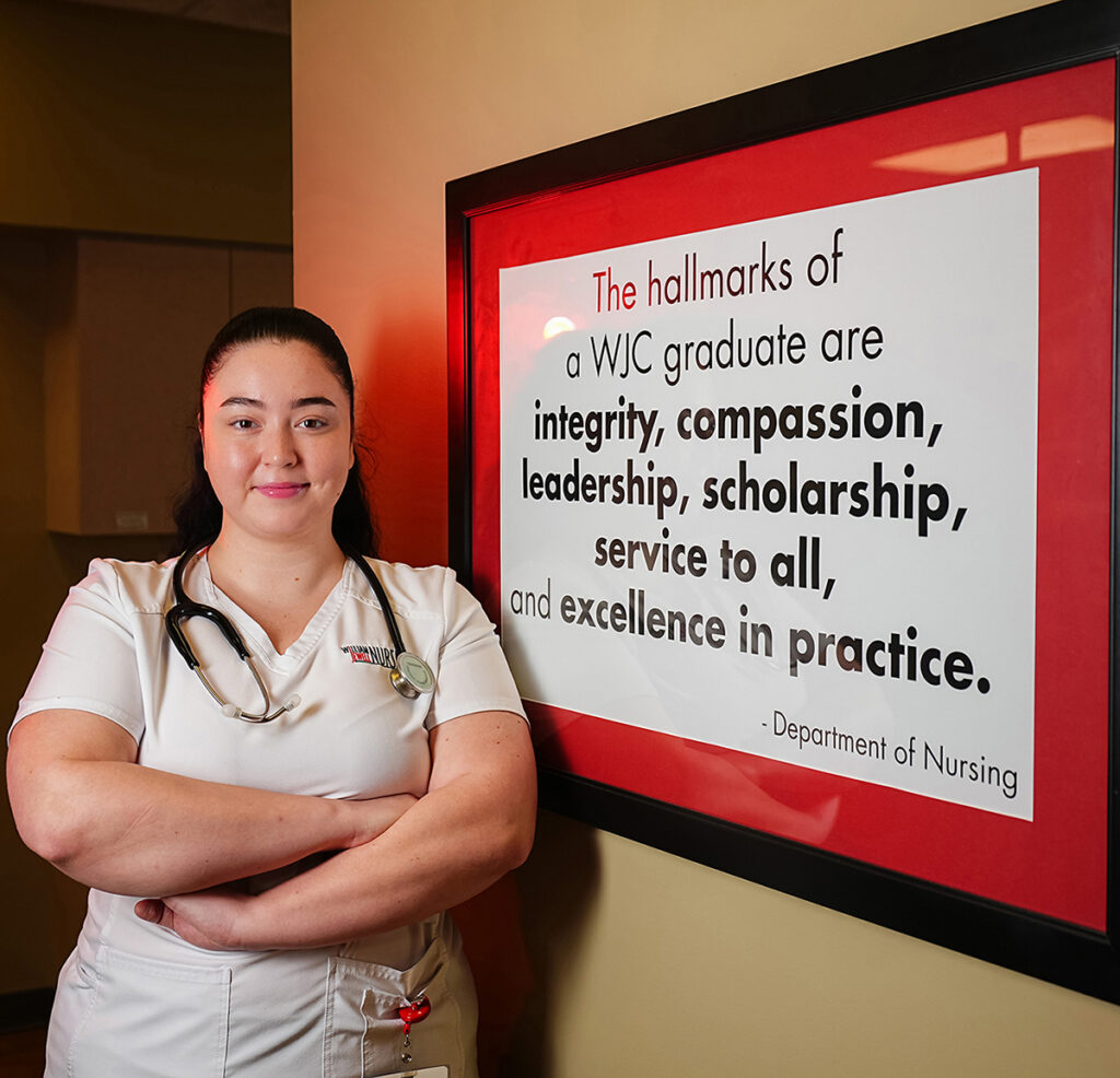 female nursing student in scrubs, wearing stethoscope, posing in nursing department by mission sign