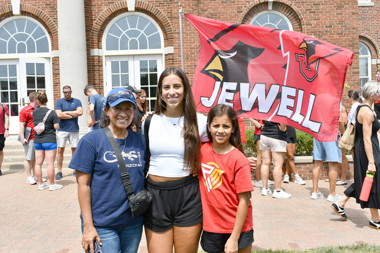 student posing with mom and sister in front of Chapel with Jewell flag in background