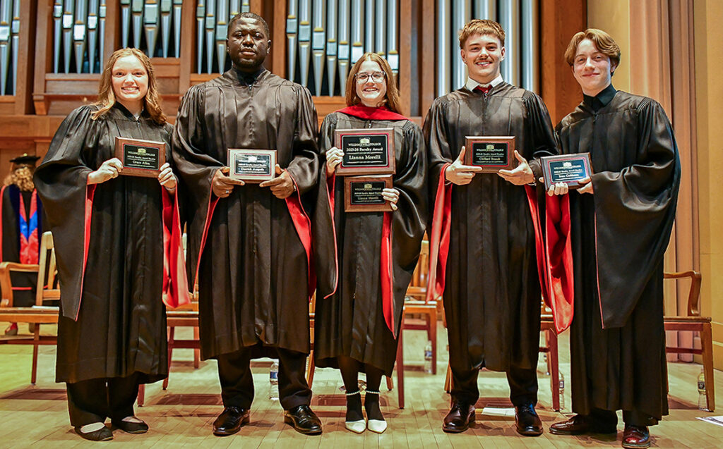 5 students in graduation gowns holding awards plaques on stage