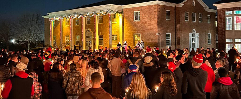 crowd holding candles in front of building with Christmas lights